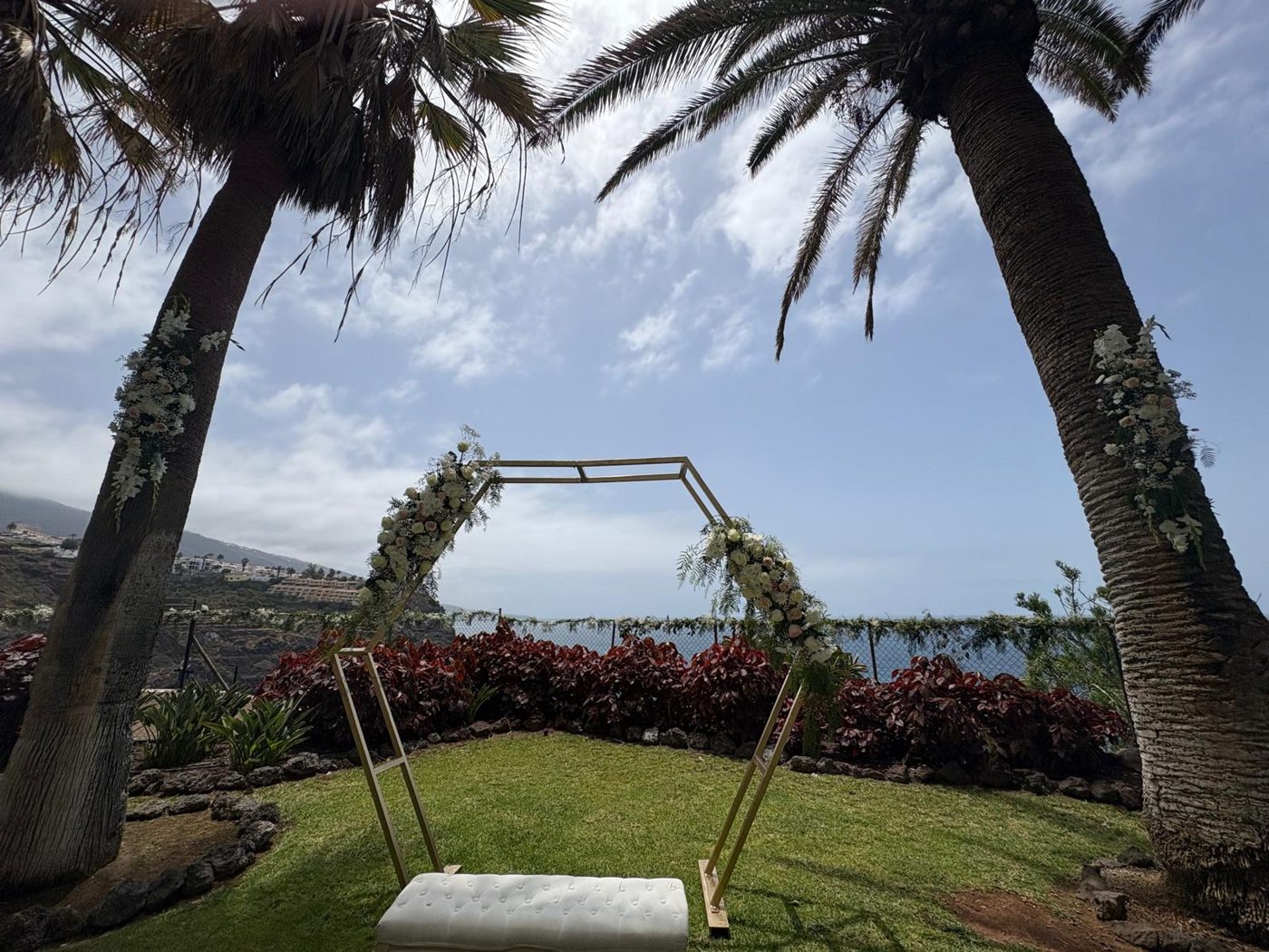 Arco hexagonal dorado con flores blancas entre palmeras y vistas al mar