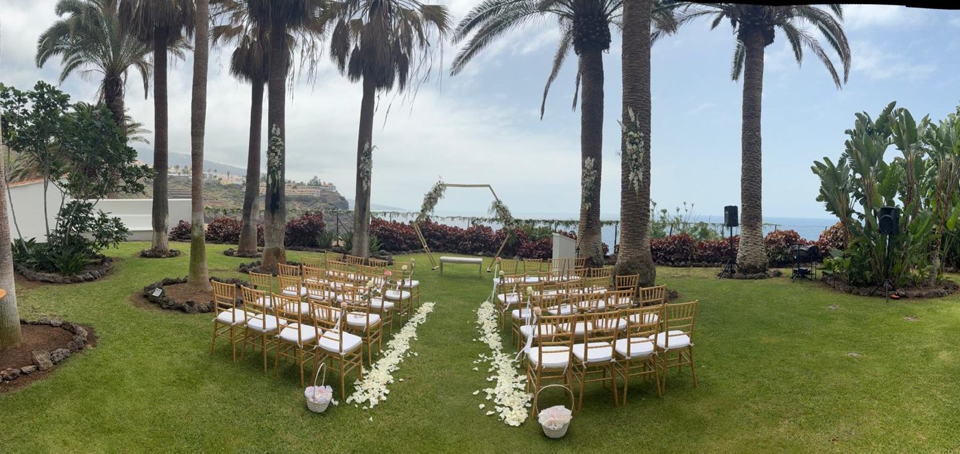 Vista panorámica de la ceremonia con sillas, arco floral, palmeras y mar