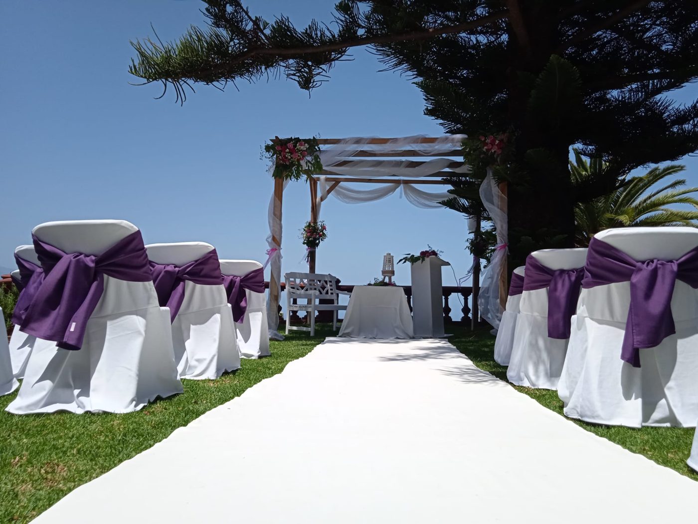 Sillas blancas con lazo morado y arco de ceremonia bajo un pino