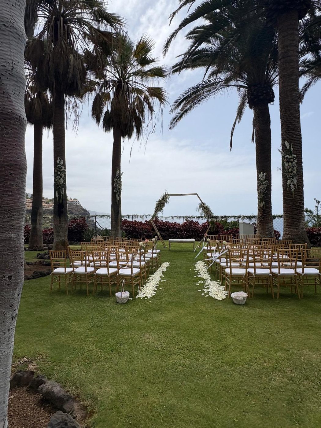 Pasillo de sillas doradas hacia el arco floral, con palmeras y vistas al mar